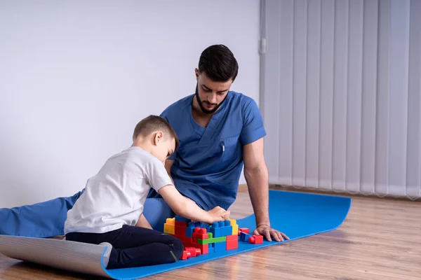 Młody lekarz z brodą w niebieskim mundurze siedzący na podłodze obok chłopca 10 lat, bawią się zabawkami edukacyjnymi, rehabilitacją koncepcyjną — Zdjęcie stockowe