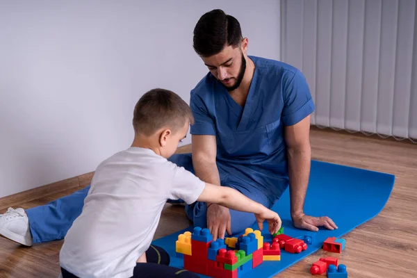 Młody lekarz z brodą w niebieskim mundurze siedzący na podłodze obok chłopca 10 lat, bawią się zabawkami edukacyjnymi, rehabilitacją koncepcyjną — Zdjęcie stockowe