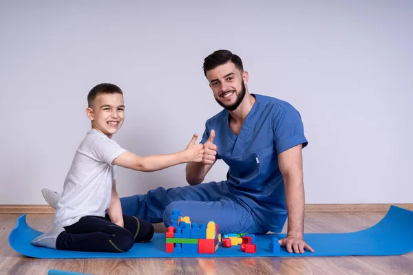 Młody lekarz z brodą w niebieskim mundurze siedzący na podłodze obok chłopca przez 10 lat, bawi się zabawkami edukacyjnymi, patrzy w kamerę i uśmiecha się — Zdjęcie stockowe