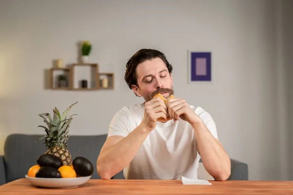 Un joven se sienta en una mesa y habla de la utilidad de las frutas, las recoge y muestra piña, aguacate, naranja. Alimento saludable —  Fotos de Stock