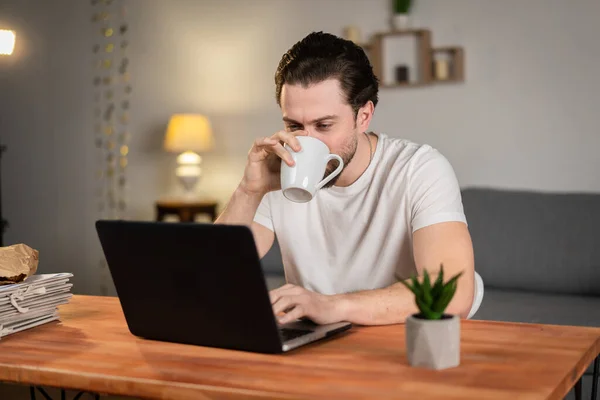 Genç bir adam evden çalışıyor ve bilgisayarla masada çay içiyor. Kötü bir fikir. Fincanda sıvı, kahve.