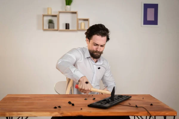 Trabalhador de escritório agressivo, homem zangado com uma barba destruir seu computador, com martelo nas mãos destruir teclado. — Fotografia de Stock