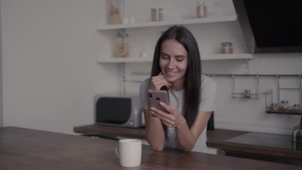 Дівчина сидить за столом на кухні, дивлячись на смартфон і інтенсивно шукаючи щось, посміхаючись, концепцію перебування вдома, купуючи онлайн — стокове відео