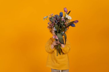 Kıvırcık saçlı şirin çocuk elinde yüzünü kaplayan çok renkli lalelerle dolu bir buket tutuyor. Sarı arka plan, tatil ve hediye kavramı.