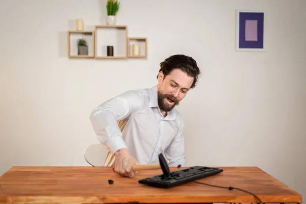 Trabalhador de escritório agressivo, homem zangado com uma barba destruir seu computador, com martelo nas mãos destruir teclado. — Fotografia de Stock