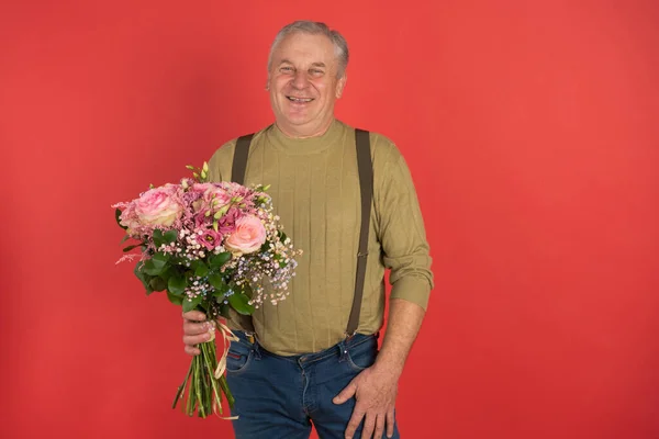 Ein älterer Mann mit einem großen schönen Blumenstrauß steht auf rotem Grund, das Konzept eines Urlaubs und eines glücklichen Alters. — Stockfoto