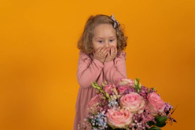 Pembe elbiseli, kıvırcık saçlı, güzel çiçeklerden oluşan bir bukete bakan ve ellerini şaşkınlıkla kapatan bir kız çocuğu. Bir tatil ve hediyeler konsepti.