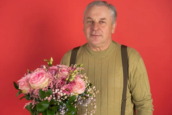 Büyük güzel bir buket çiçekle yaşlı bir adam kırmızı arka planda duruyor, tatil kavramı ve mutlu bir yaşlılık.