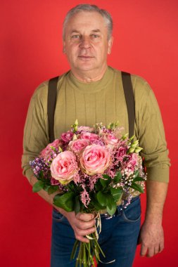 Büyük güzel bir buket çiçekle yaşlı bir adam kırmızı arka planda duruyor, tatil kavramı ve mutlu bir yaşlılık.