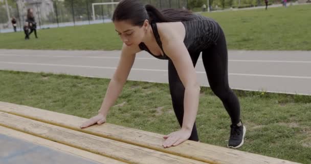 En flicka med en sport bygga driver upp på en bänk i parken, hon andas intensivt och försöker hårt, konceptet att idrotta i parken — Stockvideo