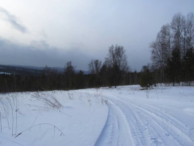 Bulutlu bir kış gününde tepenin üstündeki çayır manzarası. Dramatik bir gökyüzü. Aşağı inen yol. Kış karlı manzarası. Sibirya 'nın doğası.