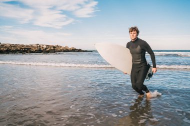 Kolunun altında sörf tahtasıyla sudan çıkan genç sörfçünün portresi. Spor ve su sporları konsepti.
