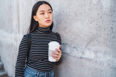 Asyalı genç bir kadının portresi beton binaya karşı kahve fincanı tutuyor..