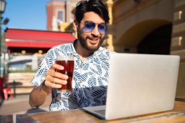 Genç yakışıklı adam yaz havasının tadını çıkarıyor ve dışarıda bira içerken bilgisayarını kullanıyor. Teknoloji ve şehir kavramı.
