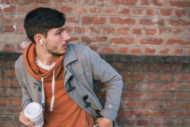 Kulaklıklı genç bir adamın portresi ve tuğla duvara dayanmış bir fincan kahve. Açık havada. Şehir konsepti.