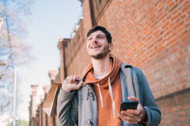 Sokakta yürüyen, alışveriş torbaları tutan ve cep telefonunu kullanan yakışıklı bir adamın portresi. Kentsel, alışveriş konsepti.