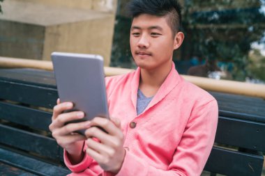 Dışarıdaki bankta otururken dijital tabletini kullanan genç Asyalı adamın portresi. Teknoloji konsepti.
