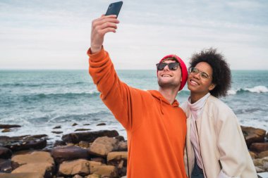 İki genç arkadaşın birlikte iyi vakit geçirdikleri ve deniz kenarında akıllı telefonlarla selfie çektikleri portre..