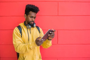 Kırmızı duvara karşı dijital tabletini ve kulaklıklarını kullanan genç afro adamın portresi. Teknoloji ve şehir kavramı.