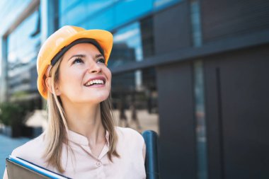 Profesyonel mimar kadın portresi sarı kask takıyor ve açık havada modern binalara bakıyor. Mühendis ve mimar kavramı.