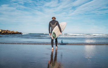 Genç sörfçü siyah sörf kıyafetiyle sörf tahtasıyla suya giriyor. Spor ve su sporları konsepti.