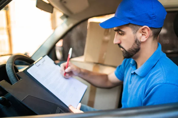 Portrait of a delivery man checking the delivery list while sitting in van. Delivery and shipping concept.