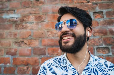 Yazlık giysiler giyen ve kulaklıkla tuğla duvara karşı müzik dinleyen genç yakışıklı adam. Şehir konsepti.