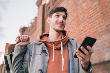 Sokakta yürüyen, alışveriş torbaları tutan ve cep telefonunu kullanan yakışıklı bir adamın portresi. Kentsel, alışveriş konsepti.