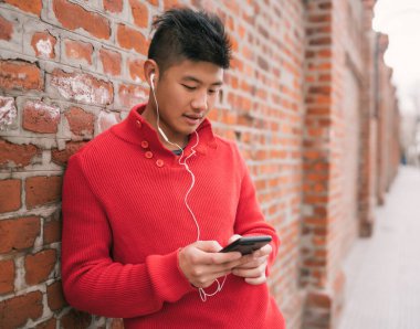 Genç Asyalı adamın portresi cep telefonuyla kulaklıklarını tuğla duvara dayamış. İletişim konsepti.