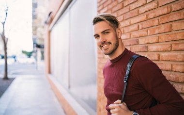 Sırt çantası taşıyan yakışıklı bir gencin portresi ve sokakta tuğladan duvara yaslanmış günlük giysiler giyiyor. Şehir konsepti.