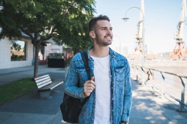 Omuzlarında sırt çantasıyla sokakta yürüyen çekici bir gencin portresi. Şehir konsepti.