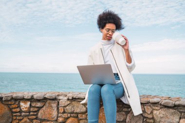 Genç Latin kadının portresi dizüstü bilgisayarını kullanıyor ve denize karşı otururken bir fincan kahve içiyor. Teknoloji konsepti.