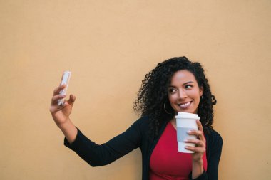 Sarı duvara bir fincan kahve tutarken mofil telefonuyla selfie çeken afro kadın portresi. Teknoloji konsepti.