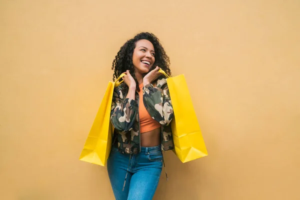 Portrait of young afro american latin woman holding shopping bags against yellow background. Shop and lifestyle concept.