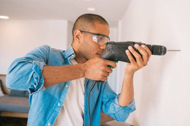 Elinde elektrikli matkapla duvarda delik açan genç Latin adamın portresi. İç tasarım ve ev yenileme konsepti.