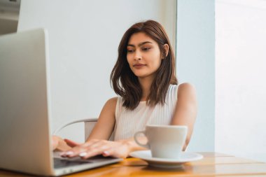 Bir kafede otururken dizüstü bilgisayarını kullanan genç Latin kadının portresi. Teknoloji ve yaşam tarzı kavramı.