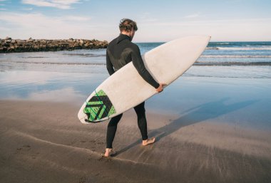 Genç sörfçü siyah sörf kıyafetiyle sörf tahtasıyla suya giriyor. Spor ve su sporları konsepti.