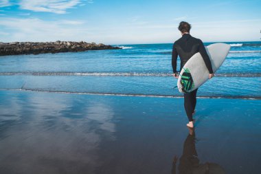 Genç sörfçü siyah sörf kıyafetiyle sörf tahtasıyla suya giriyor. Spor ve su sporları konsepti.
