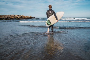 Genç sörfçü siyah sörf kıyafetiyle sörf tahtasıyla suya giriyor. Spor ve su sporları konsepti.