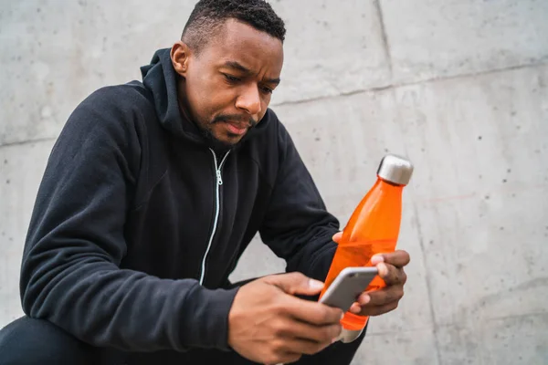 Athletic man using his mobile phone and holding a bottle of water after training against grey background. Sport and health lifestyle.