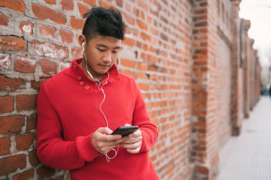Genç Asyalı adamın portresi cep telefonuyla kulaklıklarını tuğla duvara dayamış. İletişim konsepti.