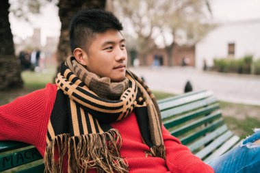 Genç Asyalı bir adamın portresi, kışlık giysiler giymiş ve rahatlamış bir şekilde parkta bir bankta oturuyor.. 