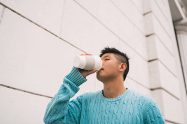 Sokakta yürürken bir fincan kahve içen genç Asyalı adamın portresi. Şehir konsepti.