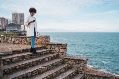 Açık havada arka planda deniz olan cep telefonunu kullanan genç ve güzel bir Latin kadının portresi. Teknoloji ve iletişim konsepti.