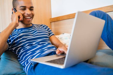 Evde dizüstü bilgisayar kullanırken kanepede uzanmış telefonda konuşan genç Latin adamın portresi. Evden işe yarıyor. Karantina, covid-19