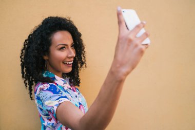 Sarı duvara karşı mofil telefonuyla selfie çeken afro kadın portresi. Teknoloji konsepti.