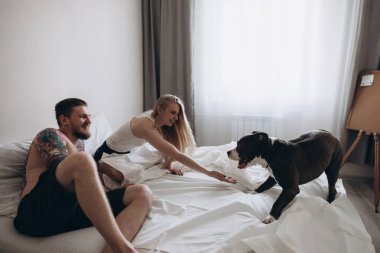 Yatağın üstüne oturan ve evde köpekle oynamayı gülümseyen çift