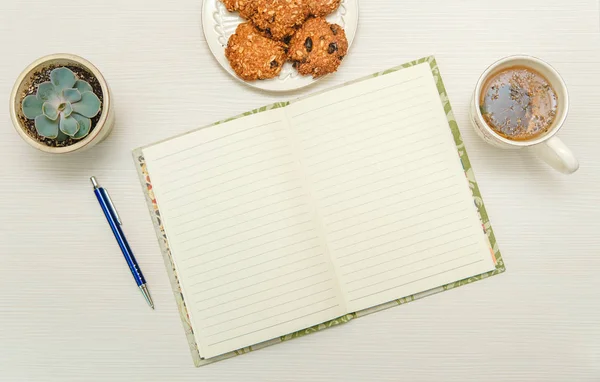 open note book with tea,cookies,plant,pen.white wooden table.