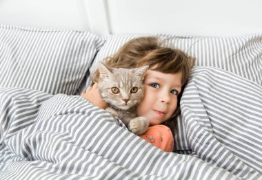 Yatakta kedisi olan çocuk portresi. Küçük kız gülümsüyor ve yatakta eğleniyor. Garip bir karantina evi izolasyonu. COVID 19