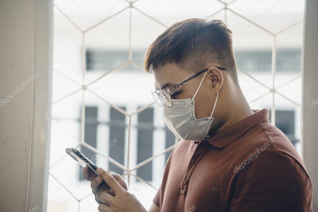 Close up of one young Asian man wearing a white surgical face mask and using a mobile phone during new type Coronavirus Covid-19 pneumonia outbreak and pm 2.5 smog air pollution crisis in big city